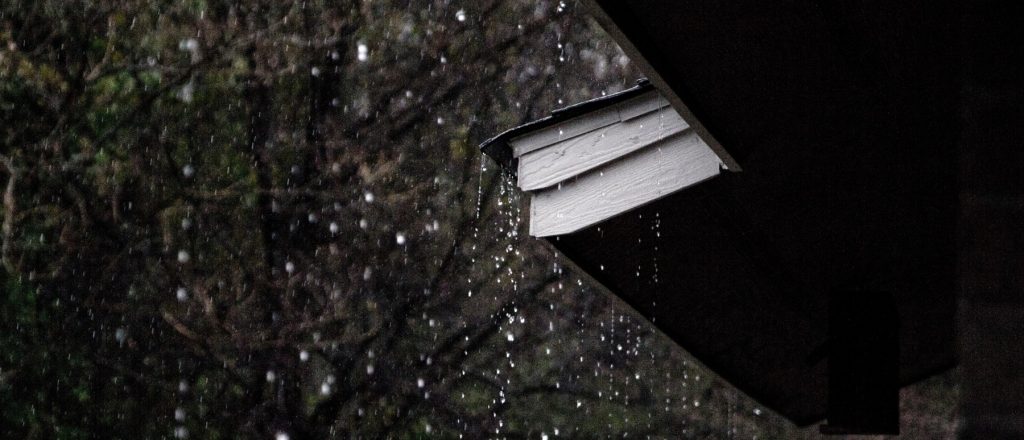 Heavy rain coming down and dripping off the corner of a roof to illustrate Rainy roof installation Warner Roofing